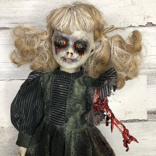 A Royally Creepy Creations doll featuring messy blonde hair, pale cracked skin, red-rimmed eyes, blood-stained cheeks, a dark dress, and a torn arm with exposed bone. The background is weathered wood.