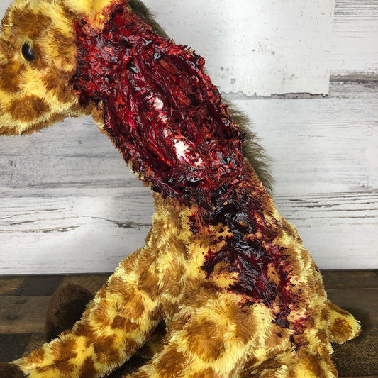 A Royally Creepy Creations plush giraffe toy features realistic special effects makeup with large, bloody wounds on its neck and shoulder, set against a wooden background.