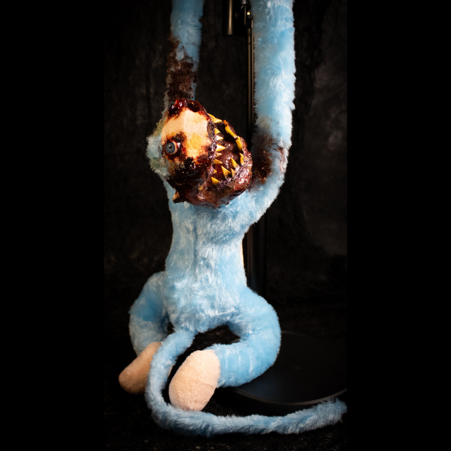 A light blue plush monkey hanging from a black stand against a dark background, with a grotesque, exposed-bone horror face and dark brown decayed details on its arms and head.