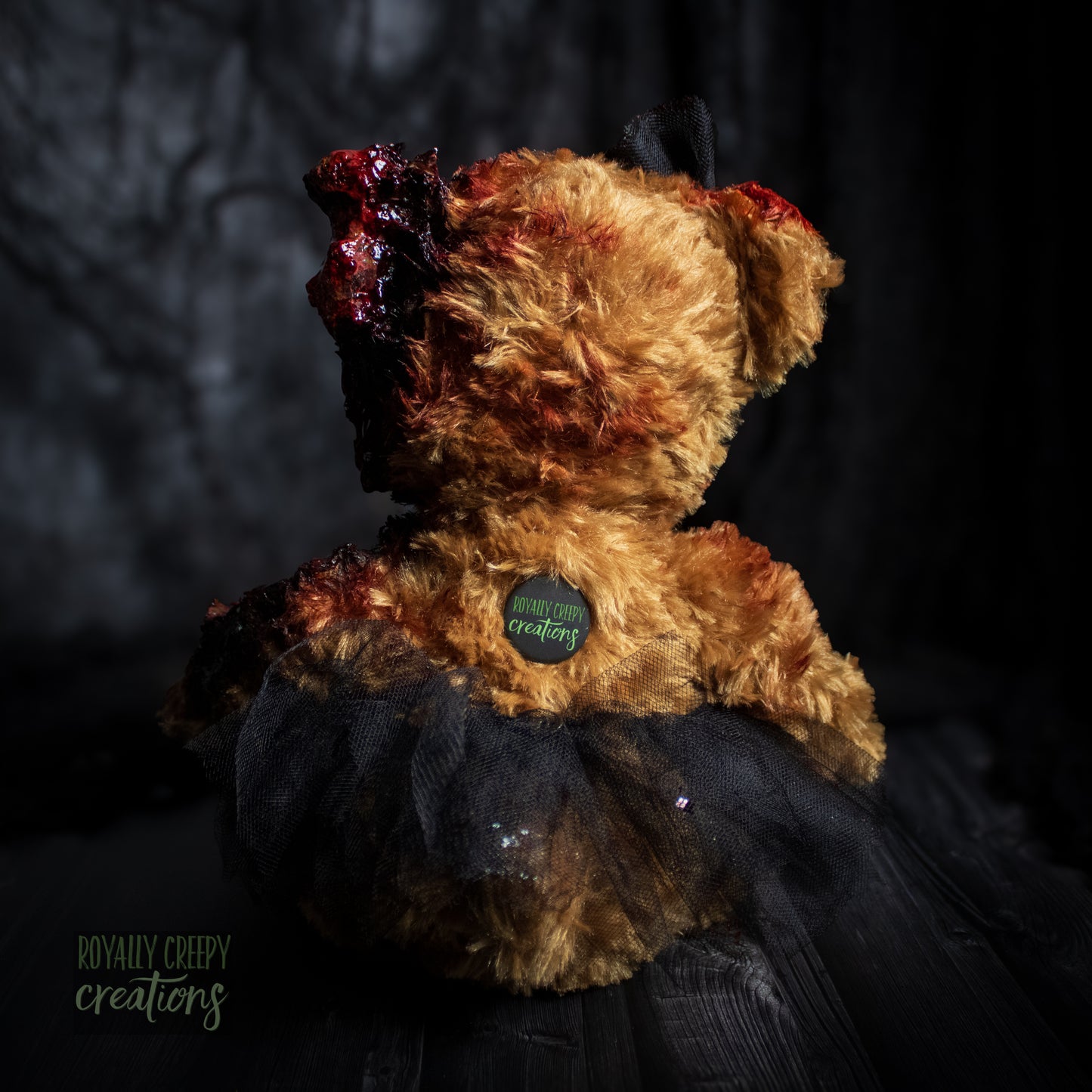 A horror-themed plush teddy bear with brown fur, bloody details on its head and shoulder, and a black tulle tutu, photographed against a dark textured background. A small circular tag on the back reads 'Royally Creepy Creations'.