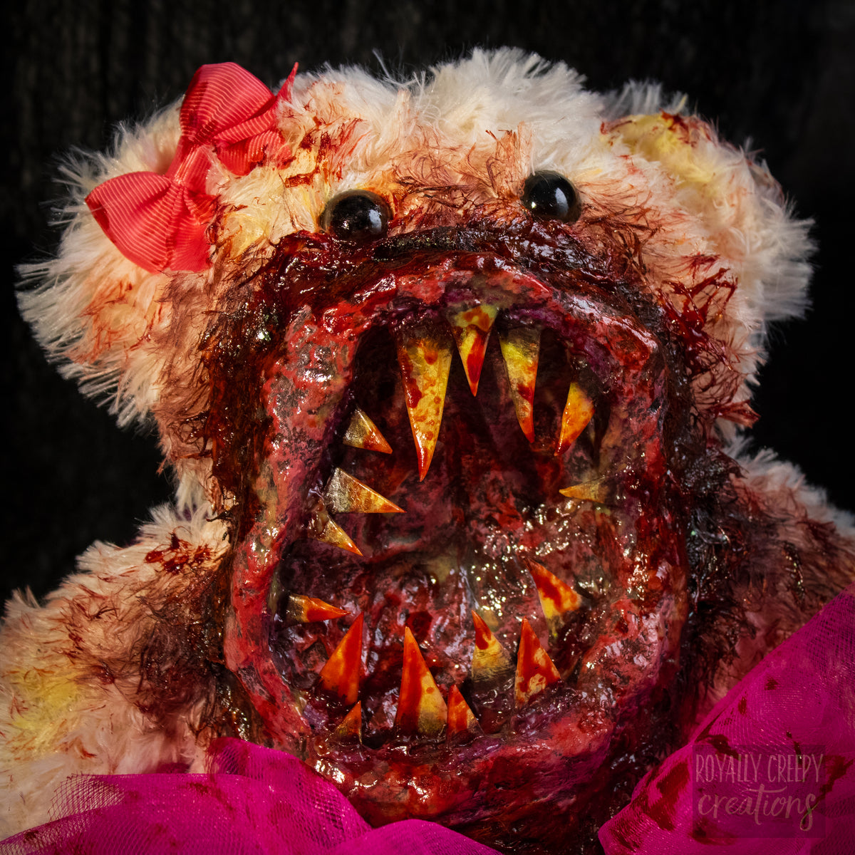A close-up of a horror-themed plush teddy bear with a bloody, grotesque mouth showing sharp teeth, a bright pink tutu, and a pink bow on its head, set against a dark background.