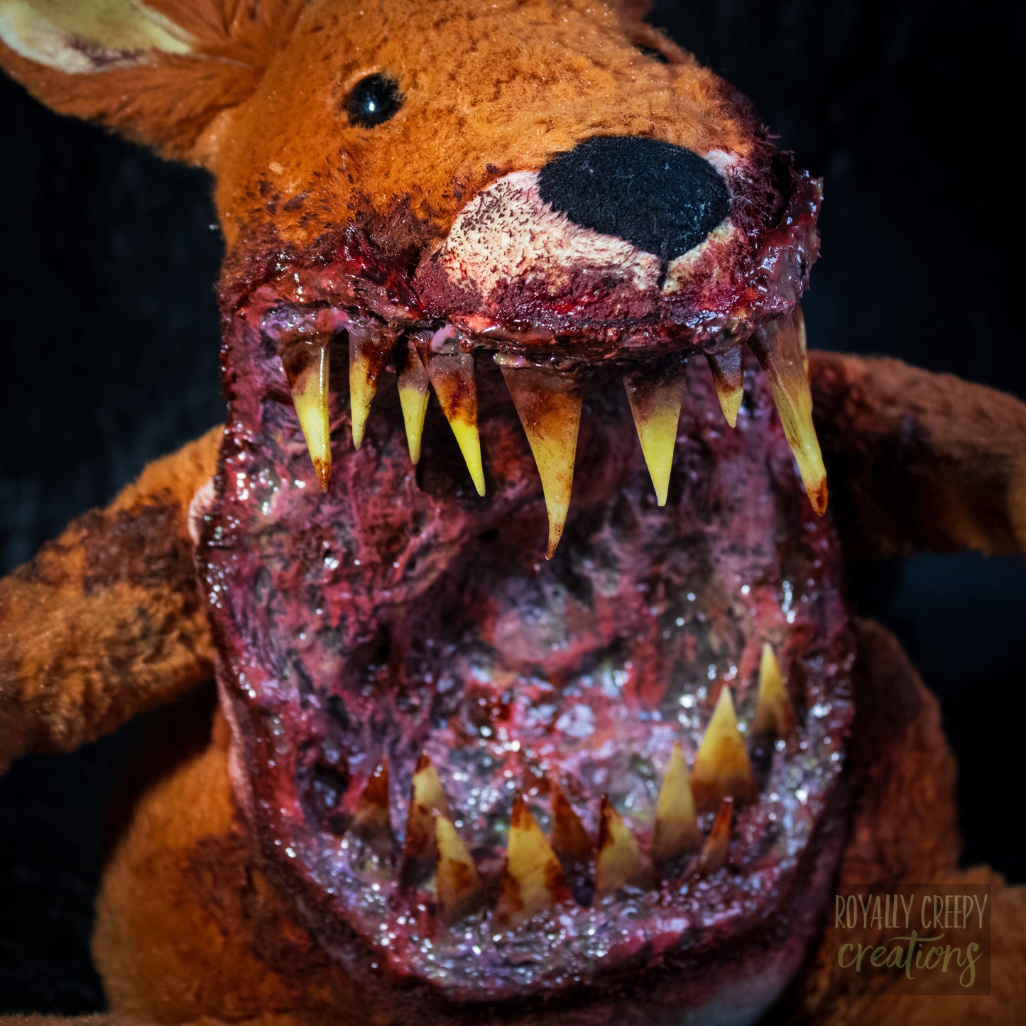 A close-up of a horror-themed plush toy with a mutilated, bloody mouth, large yellowed teeth, and a dark black nose, set against a dark background.