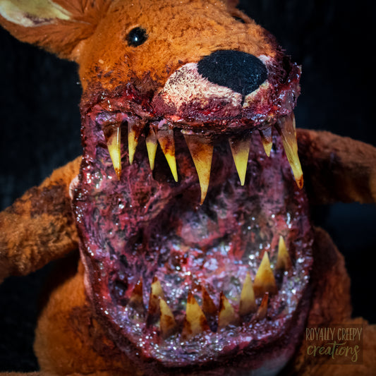 A close-up of a horror-themed plush toy with a mutilated, bloody mouth, large yellowed teeth, and a dark black nose, set against a dark background.