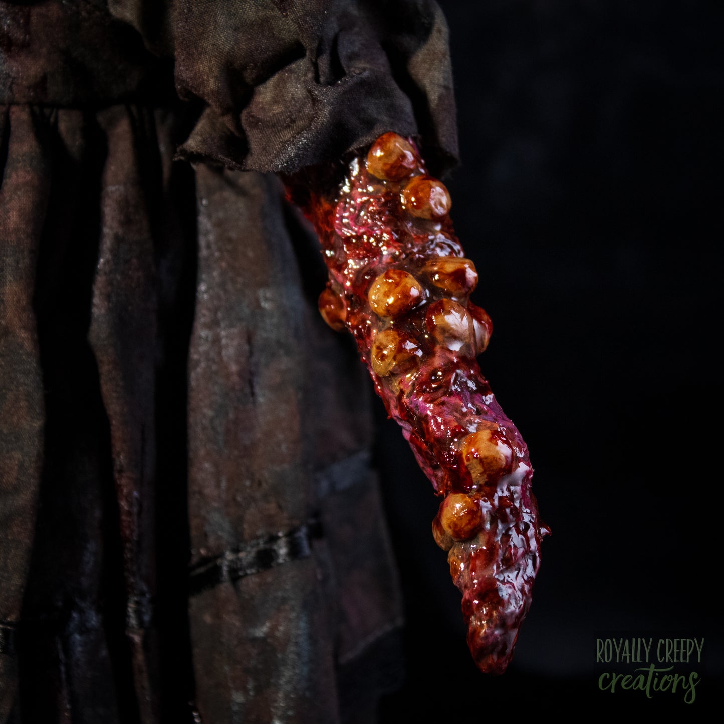 A close-up of a dark, tattered sleeve or with a mutated appendage of red tissue and raised brown bulbous growths, set against a black background.