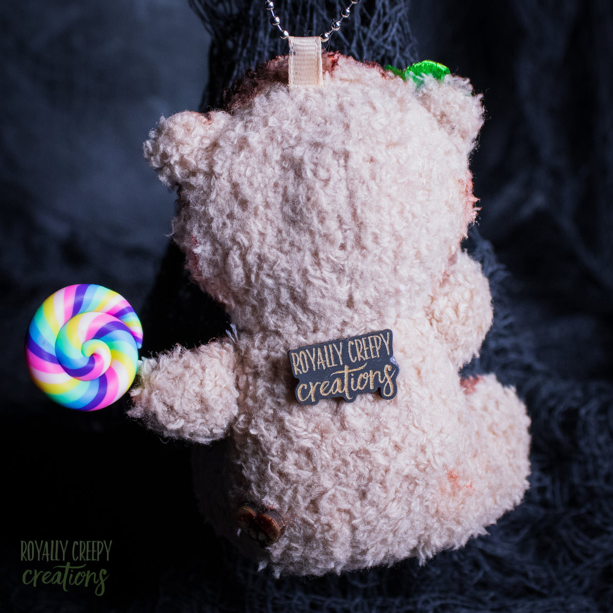 A plush teddy bear ornament or keychain in a shaggy texture, shown from the back with a colorful rainbow lollipop accessory attached on the left. The bear has a tag reading 'ROYALLY CREEPY creations' on its back, photographed against a dark textured background.