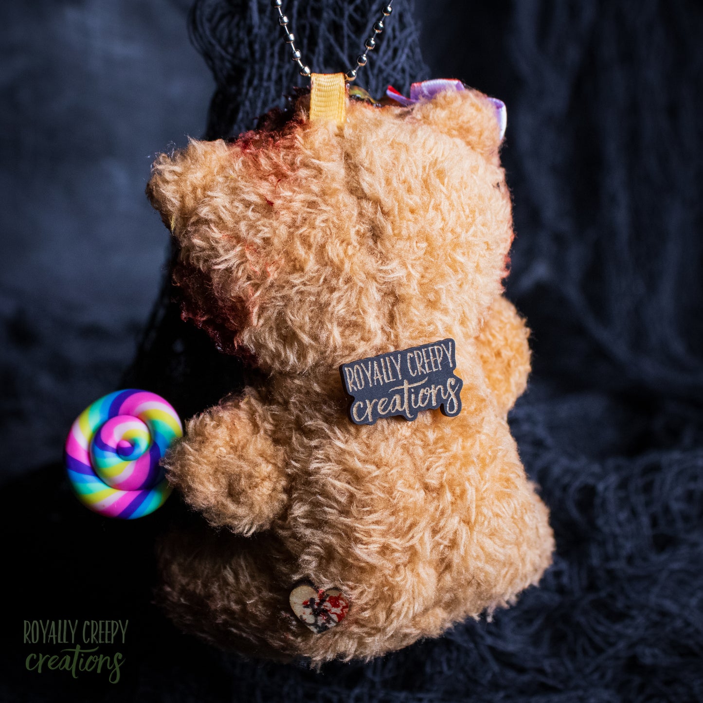A plush teddy bear ornament in light brown fur, holding a rainbow spiral lollipop and wearing a black patch that reads 'ROYALLY CREEPY creations'. The ornament hangs from a silver chain against a dark textured background.