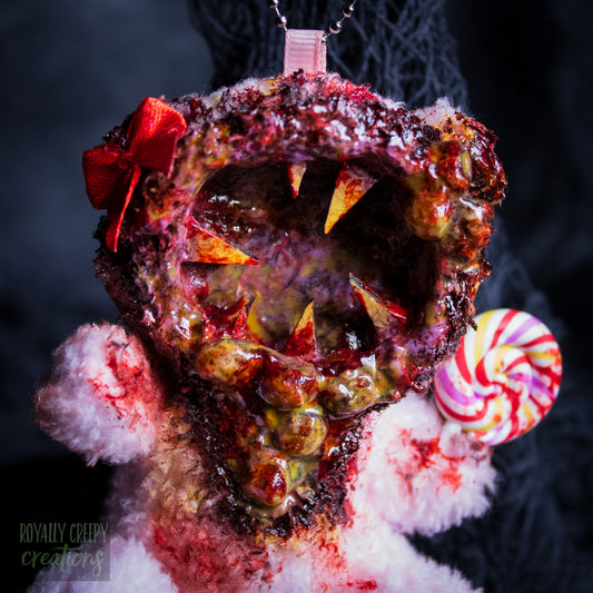 A close-up of a horror-themed plush bear keychain with a bloody, gory mouth design, fuzzy body, and lollipop accessory, shown against a black netted backdrop.