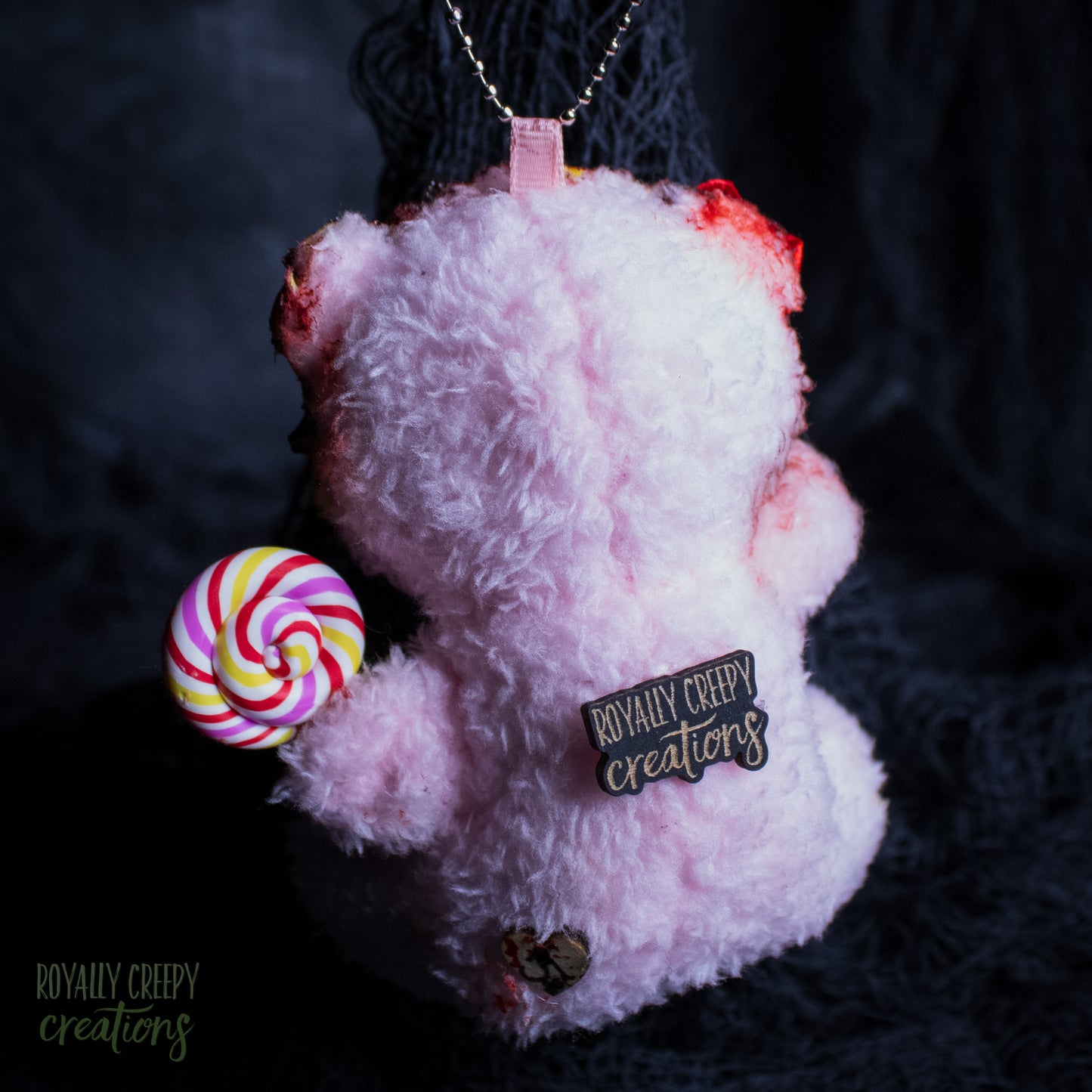 A pink plush teddy bear keychain or pendant with horror-themed details, including fake blood on the ears, a pink and white striped lollipop, and a black enamel pin reading 'Royally Creepy Creations'.