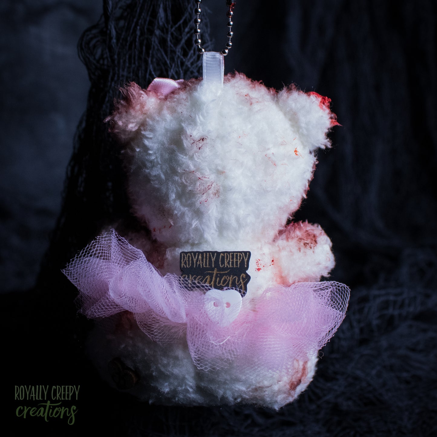 The back of a teddy bear ornament with white fluffy fur, pink tulle skirt, and fake blood splatters, featuring a 'Royally Creepy Creations' brand tag.
