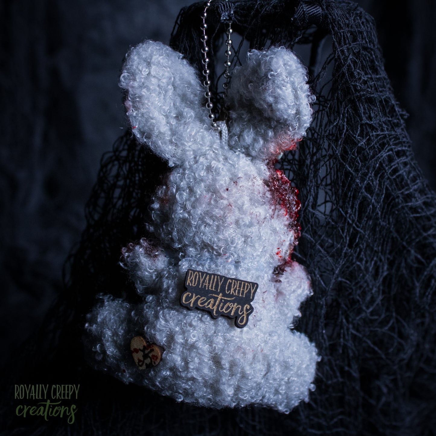 A white fluffy plush bunny keychain or bag charm with red blood-like splatter details, a metal chain, and a wooden brand tag reading 'Royally Creepy Creations,' shown against a dark netting backdrop.