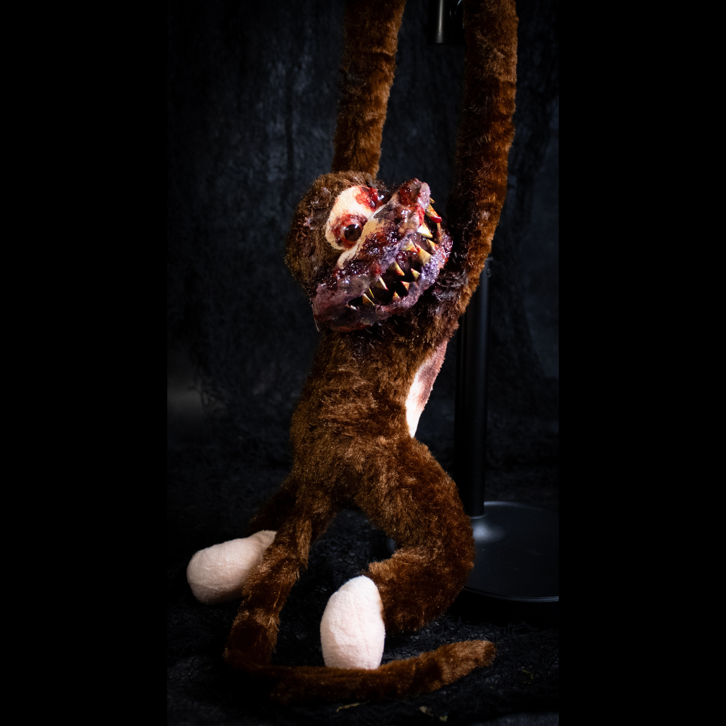 A horror-themed plush monkey toy with brown fur, pale pinkish-white feet and hands, and a grotesque bloody mouth with sharp teeth, posed against a dark textured backdrop.