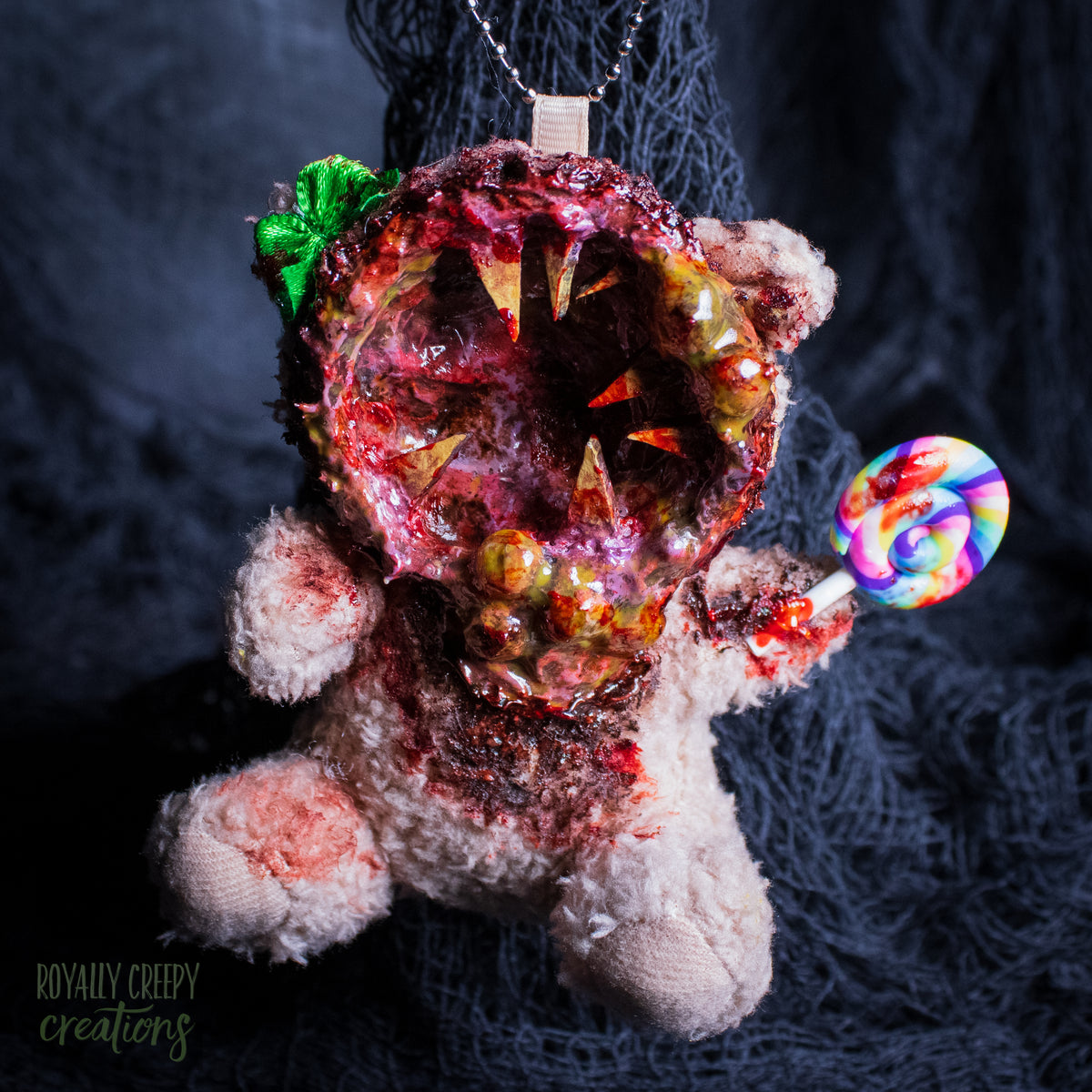 A horror-themed plush bear keychain with a bloody, gory mouth design, fuzzy body, and lollipop accessory, shown against a black netted backdrop.
