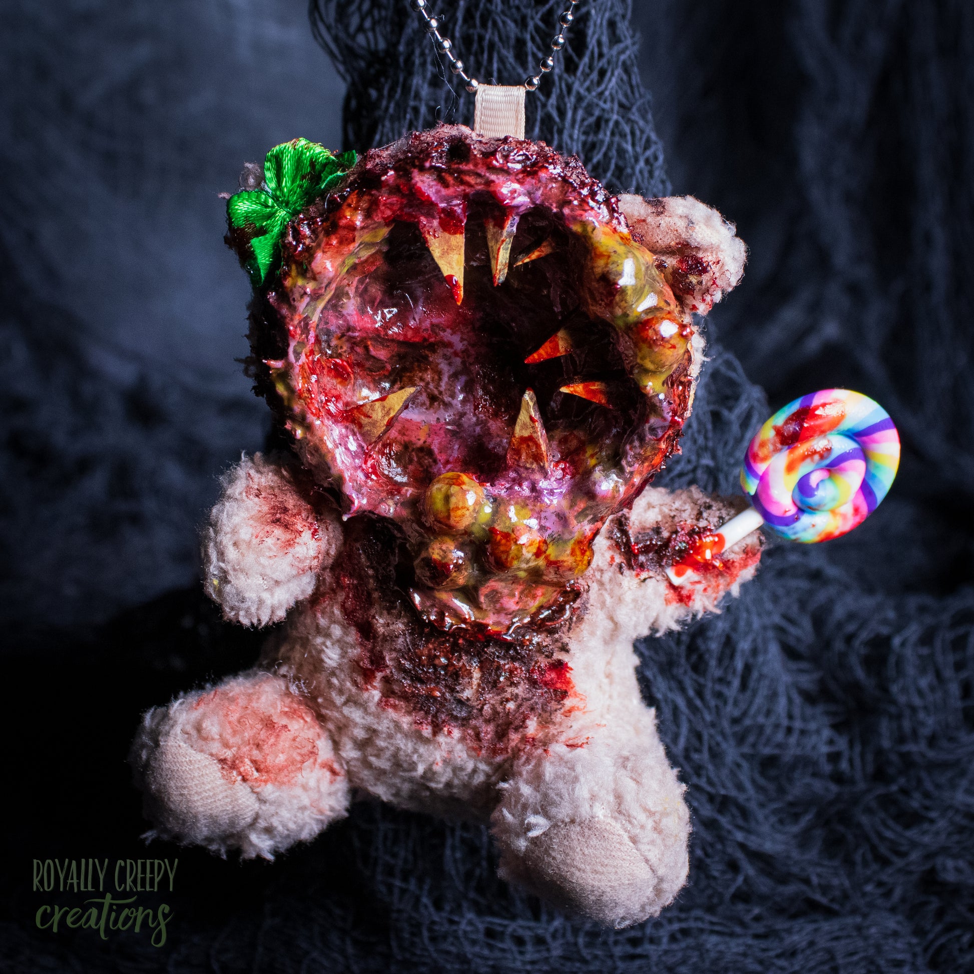 A horror-themed plush bear keychain with a bloody, gory mouth design, fuzzy body, and lollipop accessory, shown against a black netted backdrop.
