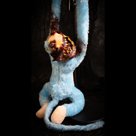 A light blue plush monkey hanging from a black stand against a dark background, with a grotesque, exposed-bone horror face and dark brown decayed details on its arms and head.