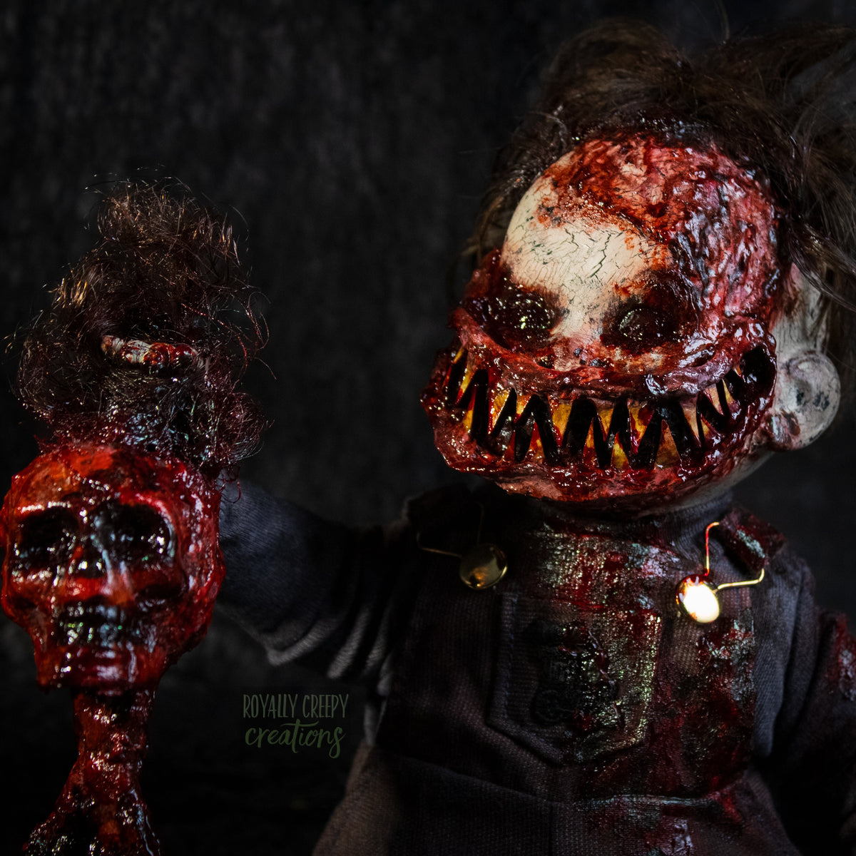 A close-up of a creepy, horror-themed doll with a big smile full of sharp teeth and a blood-splattered face, dark tattered hair, and a severed skull in it's hand, posed against a dark textured background.