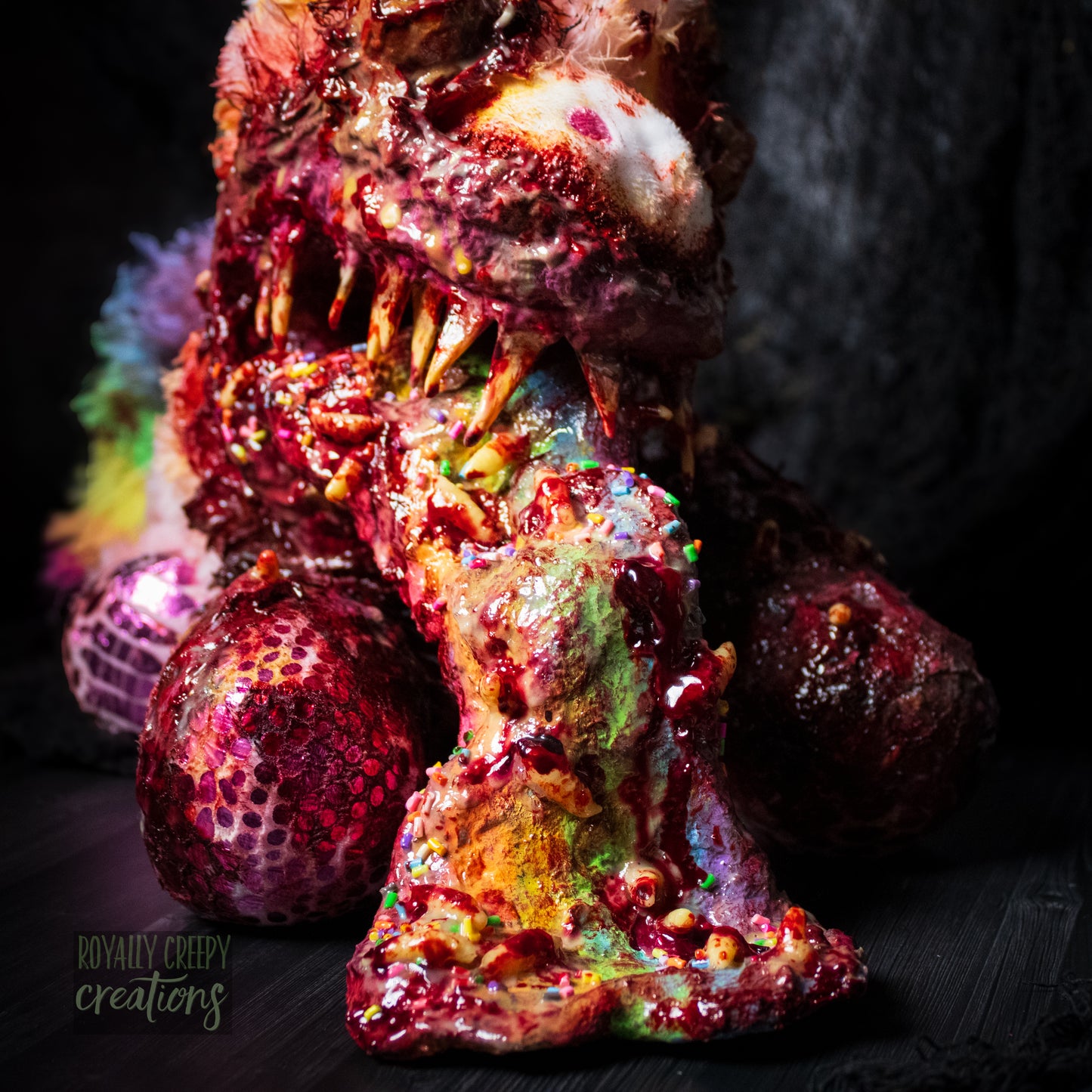Close-up front view of horror themed unicorn plush with sharp teeth vomiting a rainbow covered with maggots and colorful sprinkles and fake blood.