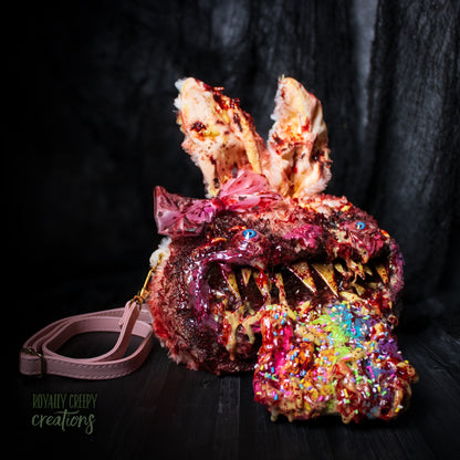 A horror-themed bunny head crossbody bag with sharp teeth, colorful sprinkles on the lower part, and a pink bow, shown with a matching pink strap on a dark background.