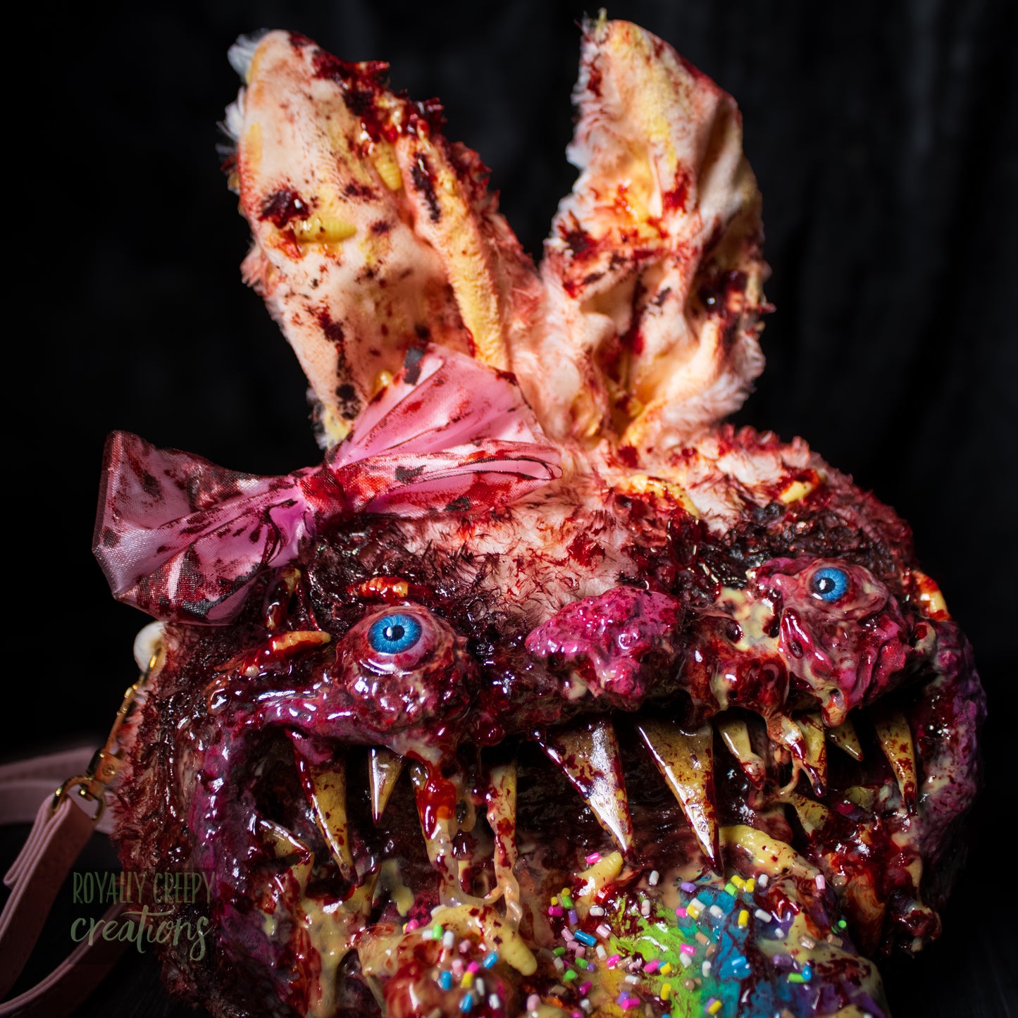 Close-up of a horror-themed bunny purse featuring a monstrous face with sharp teeth, blue eyes, a pink bow, and a colorful tongue covered in sprinkles, set against a dark background.