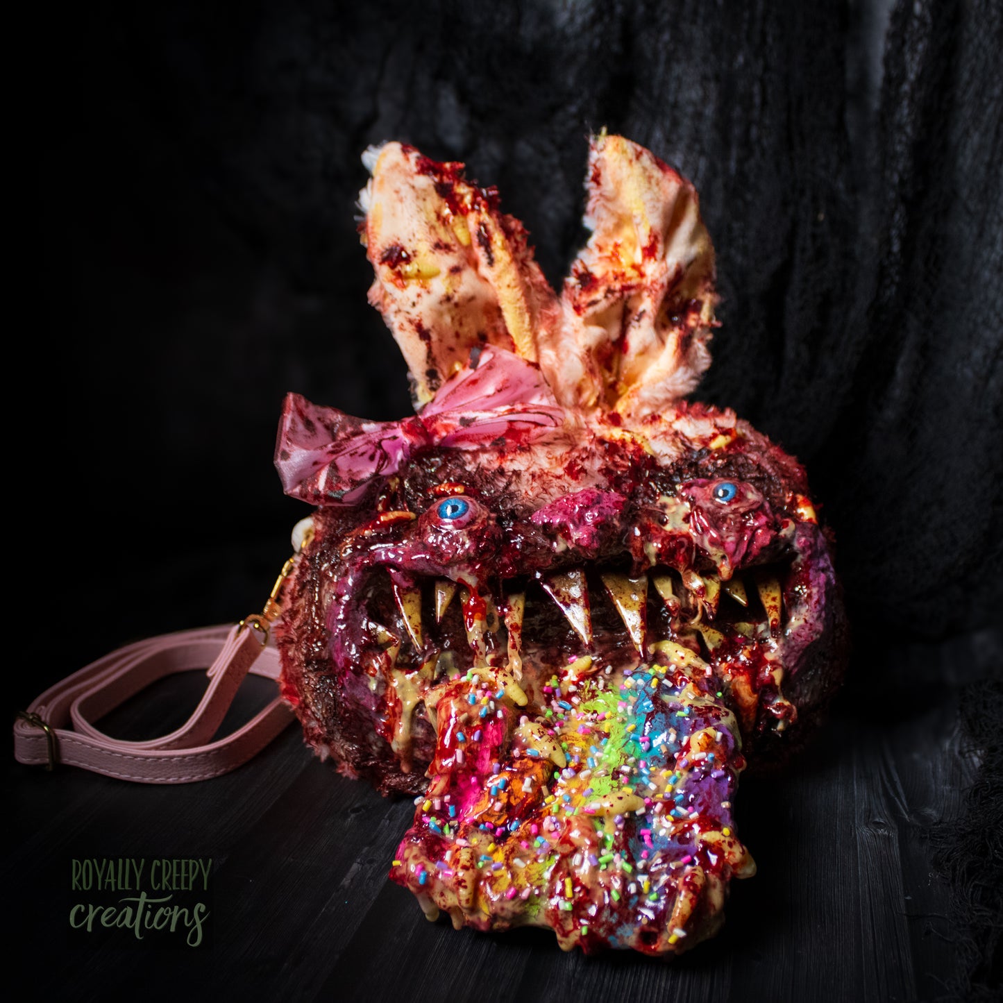 A horror-themed bunny purse featuring a monstrous face with sharp teeth, blue eyes, a pink bow, and a colorful tongue covered in sprinkles, set against a dark background.