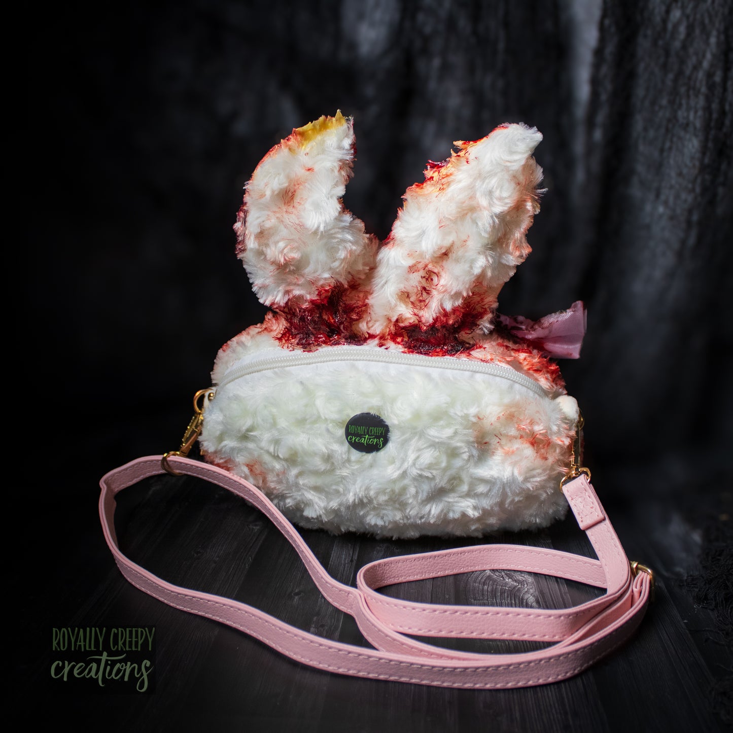 A plush bunny-shaped crossbody bag with white faux fur, red blood-like splatter details, a pink ribbon, and a long pink strap, photographed on a dark studio background.