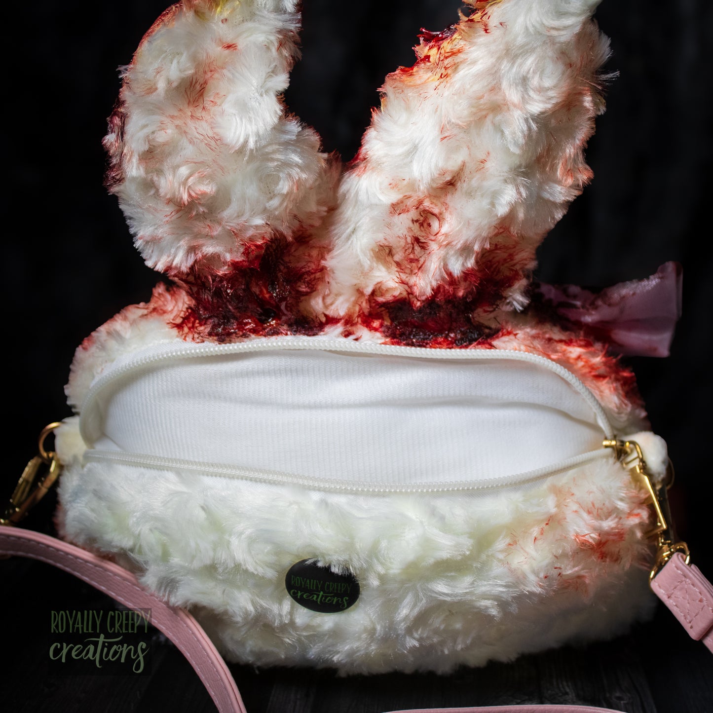 A white faux fur bunny-shaped clutch bag with red blood splatter details, a white zipper closure, gold-tone hardware, and a pink strap, shown against a dark background.