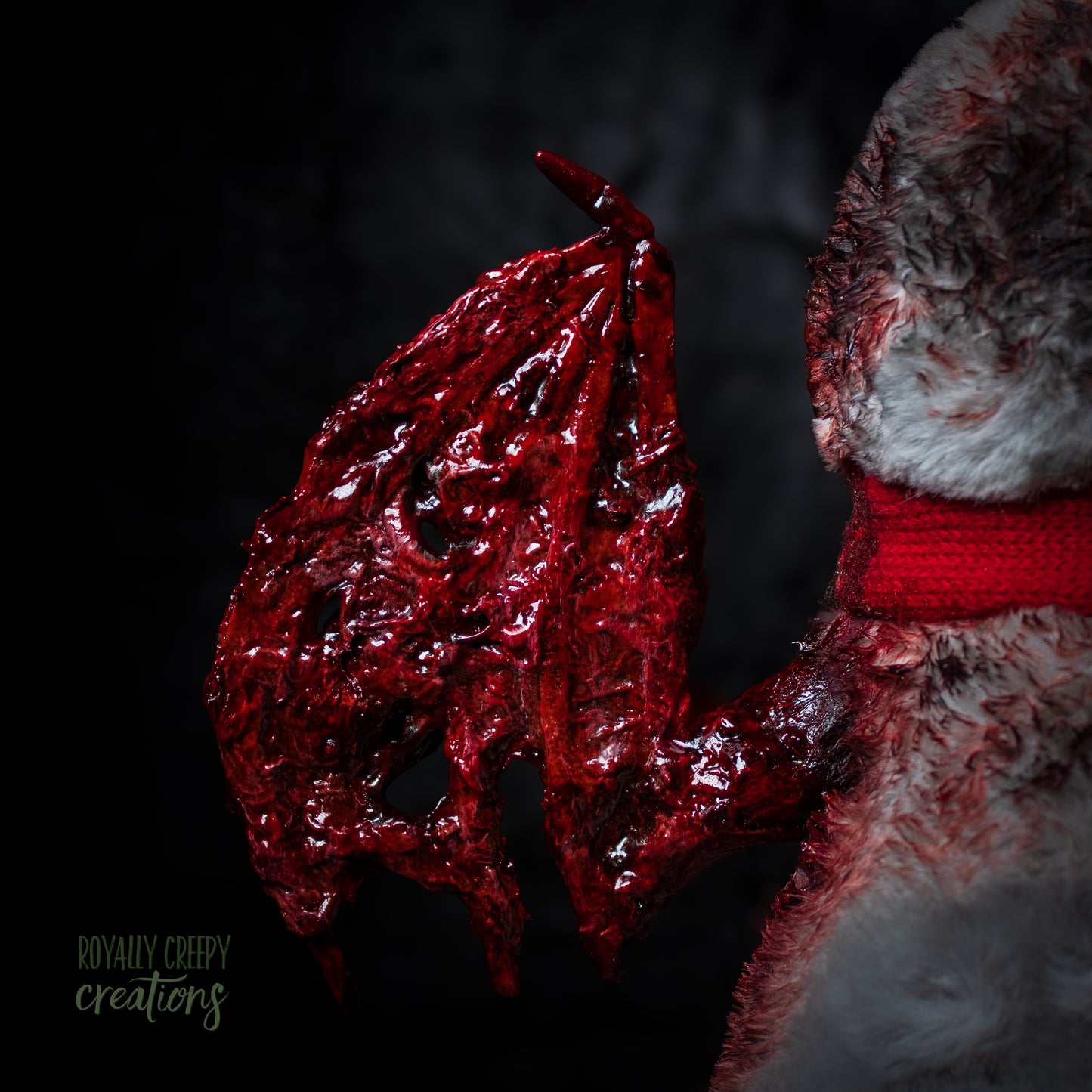 Close-up of a red textured bloody left wing attached to plush penguin with a red scarf on a dark background.