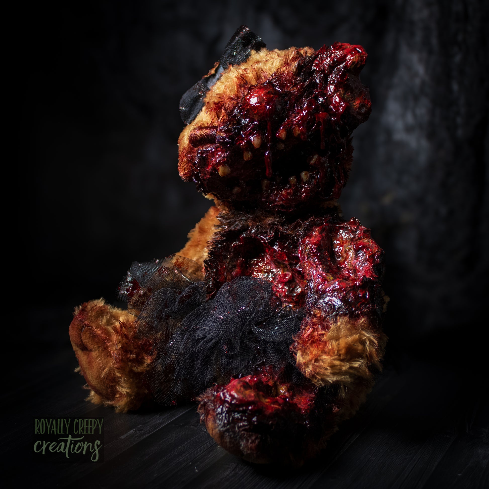 A horror-themed plush teddy bear with a grotesque, gory zombie-like face, bared teeth, and exposed flesh details, wearing a black tutu and black bow, set against a dark, moody background.