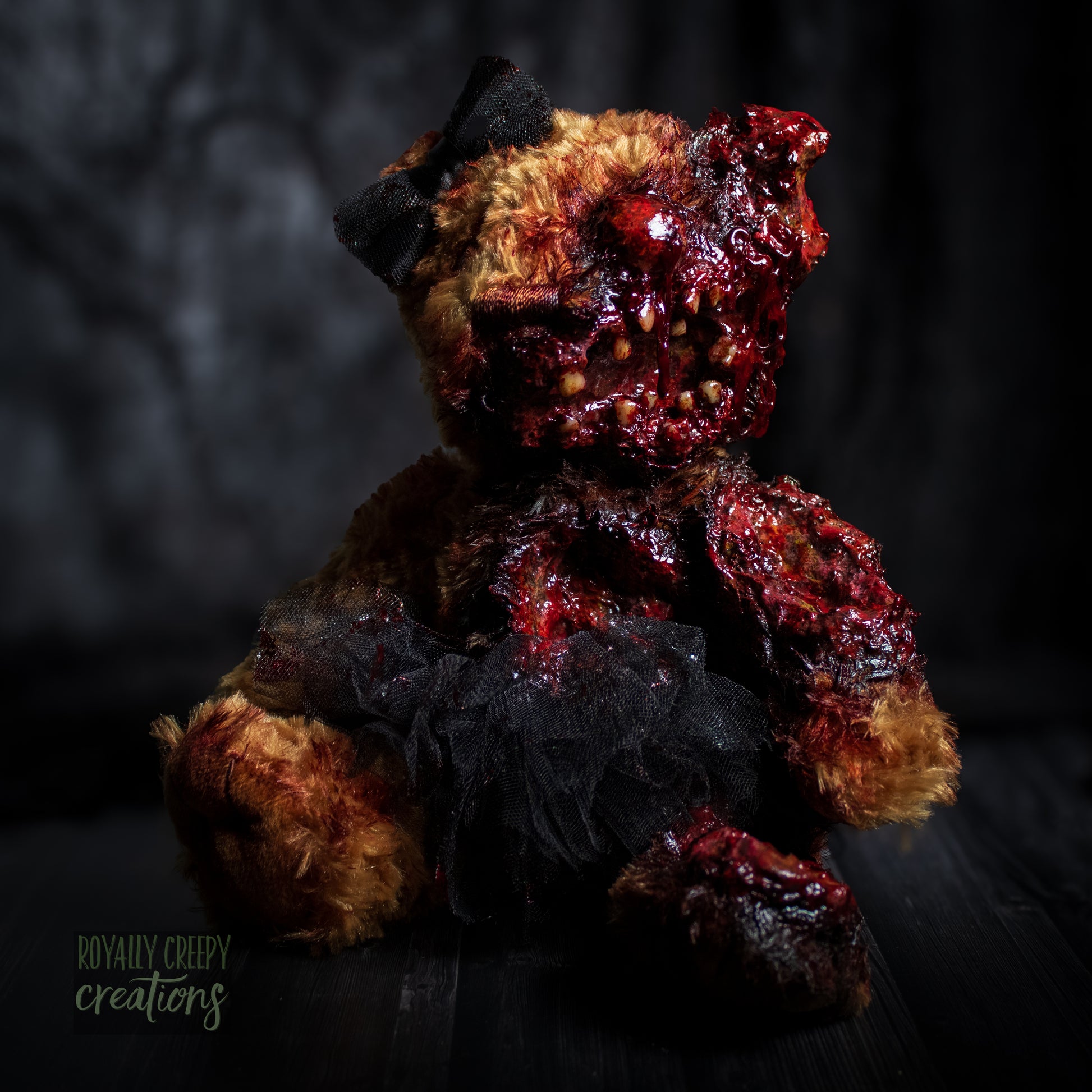 A horror-themed plush teddy bear with a grotesque, gory zombie-like face, bared teeth, and exposed flesh details, wearing a black tutu and black bow, set against a dark, moody background.