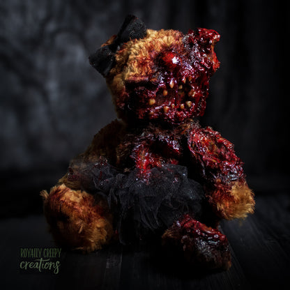 A horror-themed plush teddy bear with a grotesque, gory zombie-like face, bared teeth, and exposed flesh details, wearing a black tutu and black bow, set against a dark, moody background.