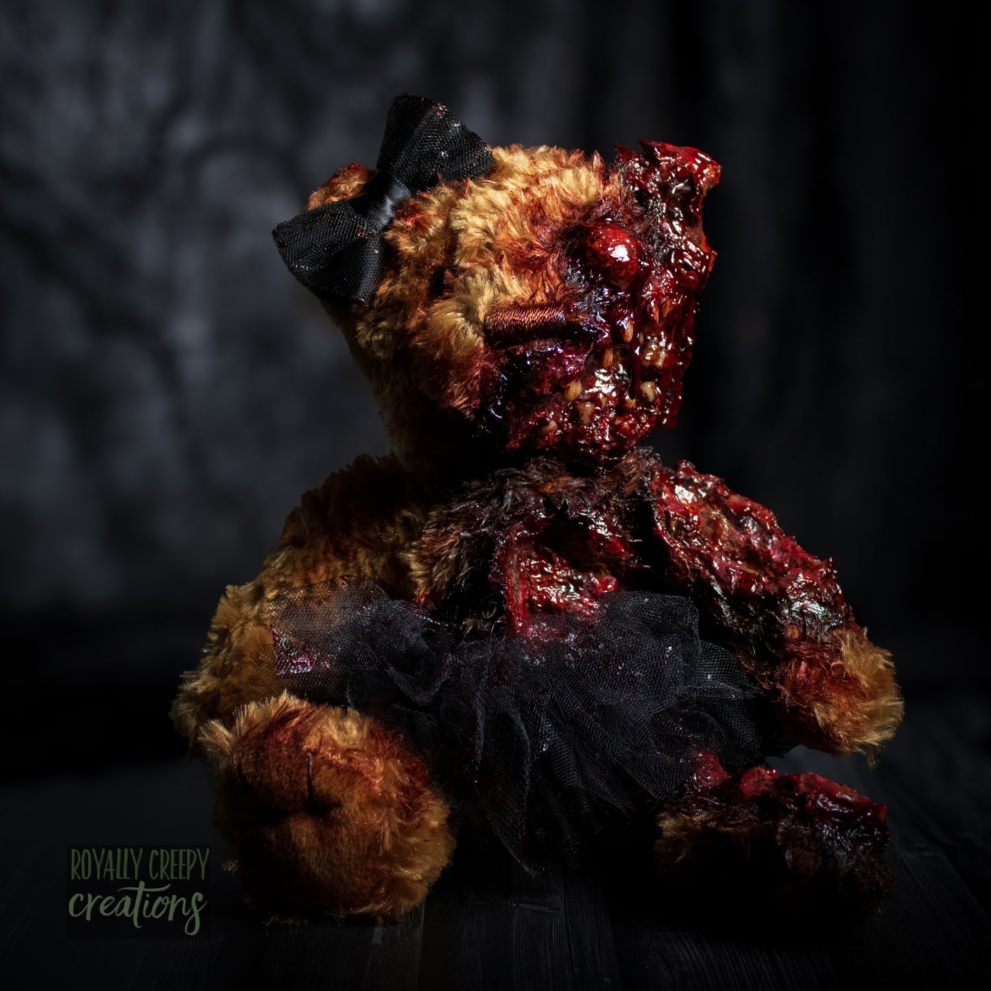 A horror-themed plush teddy bear with a grotesque, gory zombie-like face, bared teeth, and exposed flesh details, wearing a black tutu and black bow, set against a dark, moody background.