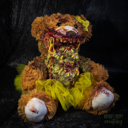 A horror-themed plush teddy bear with a bloody mangled mouth, exposed fake vomit, and a bright yellow tutu and bow, posed against a dark textured backdrop.