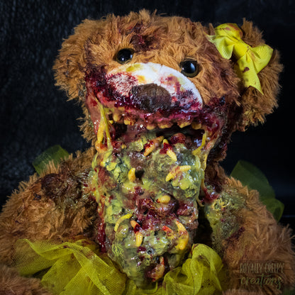 Close-up of a horror-themed plush teddy bear with a bloody mangled mouth, exposed fake vomit, and a bright yellow tutu and bow, posed against a dark textured backdrop.