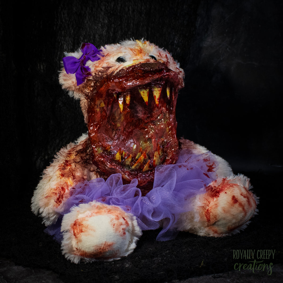 A horror-themed plush teddy bear with a grotesque, bloody mouth, sharp teeth, a purple bow, and a purple tutu on a black background.