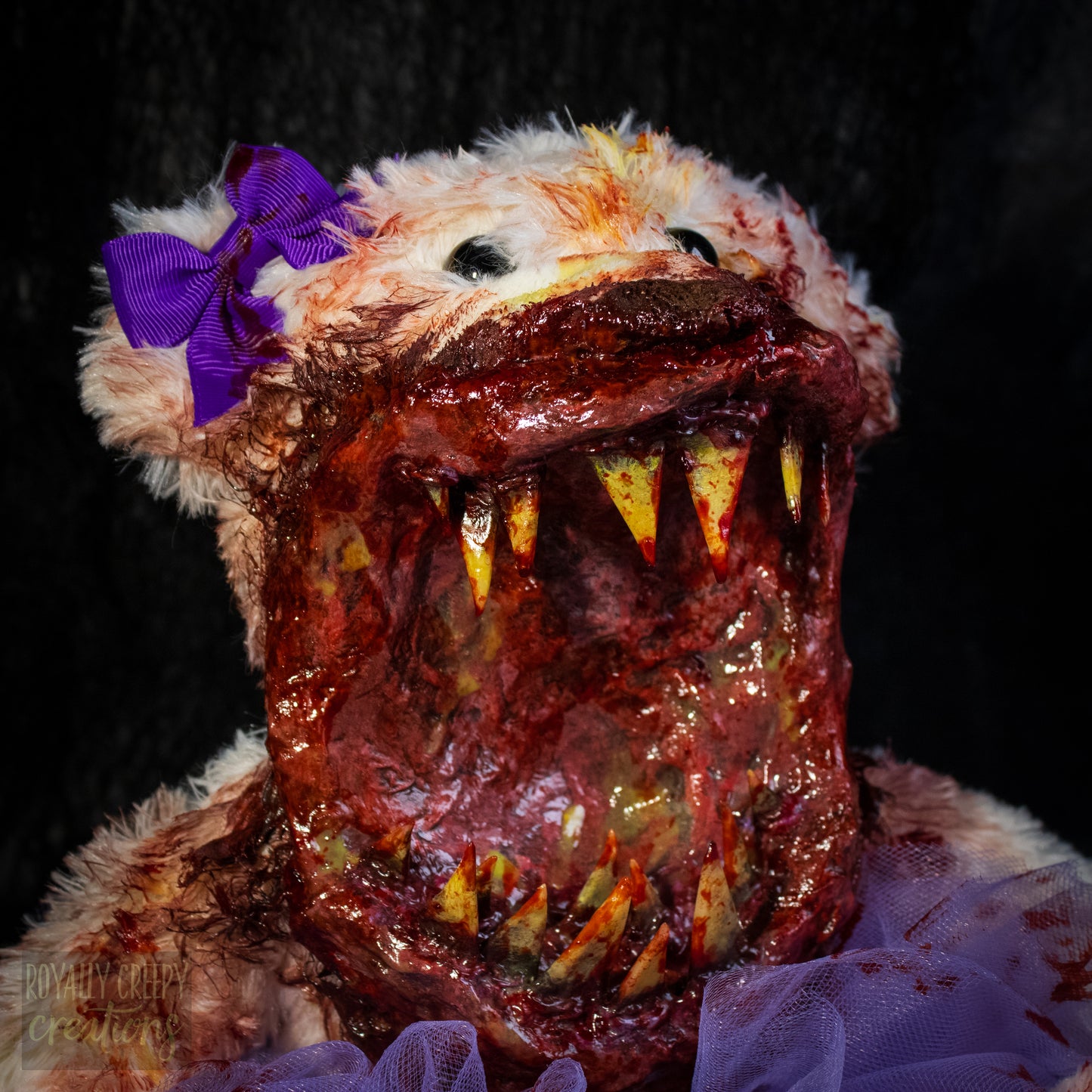 A close-up of a horror-themed plush bear with a grotesque, mouth-covered face, sharp yellowed teeth, a purple bow on its ear, and a purple tulle skirt against a black background.