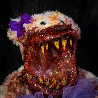 A close-up of a horror-themed plush bear with a grotesque, mouth-covered face, sharp yellowed teeth, a purple bow on its ear, and a purple tulle skirt against a black background.