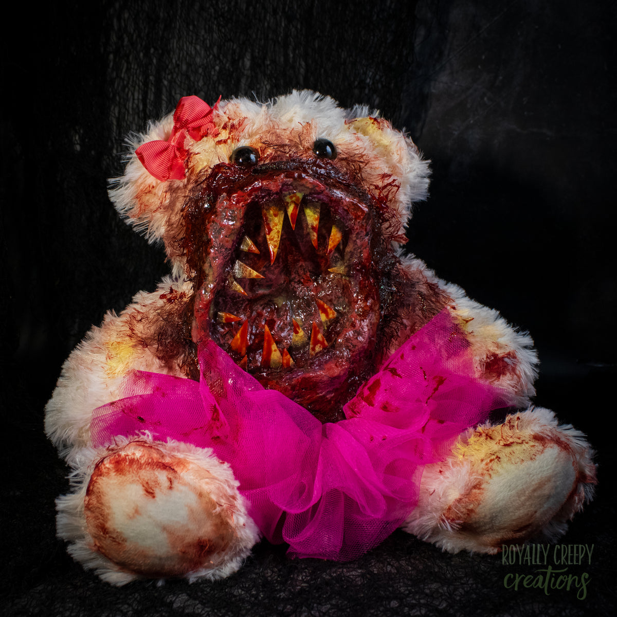 A horror-themed plush teddy bear with a bloody, grotesque mouth showing sharp teeth, a bright pink tutu, and a pink bow on its head, set against a dark background.