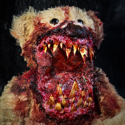 A close-up of a horror-themed plush teddy bear with a grotesque, bloody mouth showing sharp yellowed teeth, set against a dark textured background.