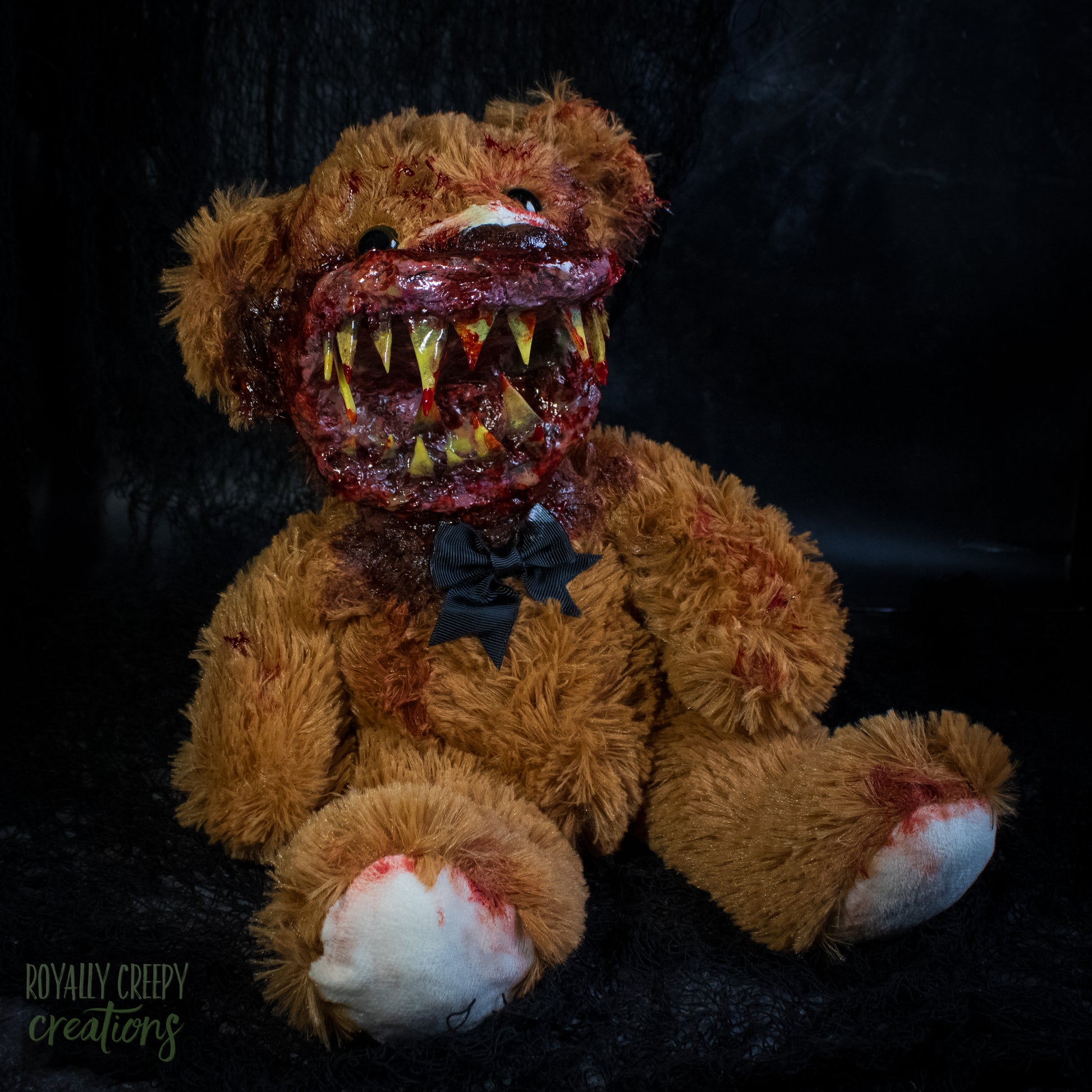 A horror-themed plush teddy bear with a grotesque bloody mouth showing yellowed teeth, sitting against a dark background. 