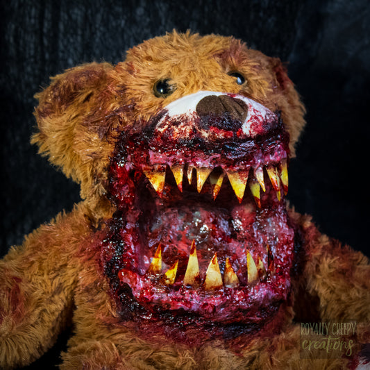 a close-ip of a horror-themed plush teddy bear with a grotesque, bloody open mouth showing sharp yellow teeth, set against a dark textured background.