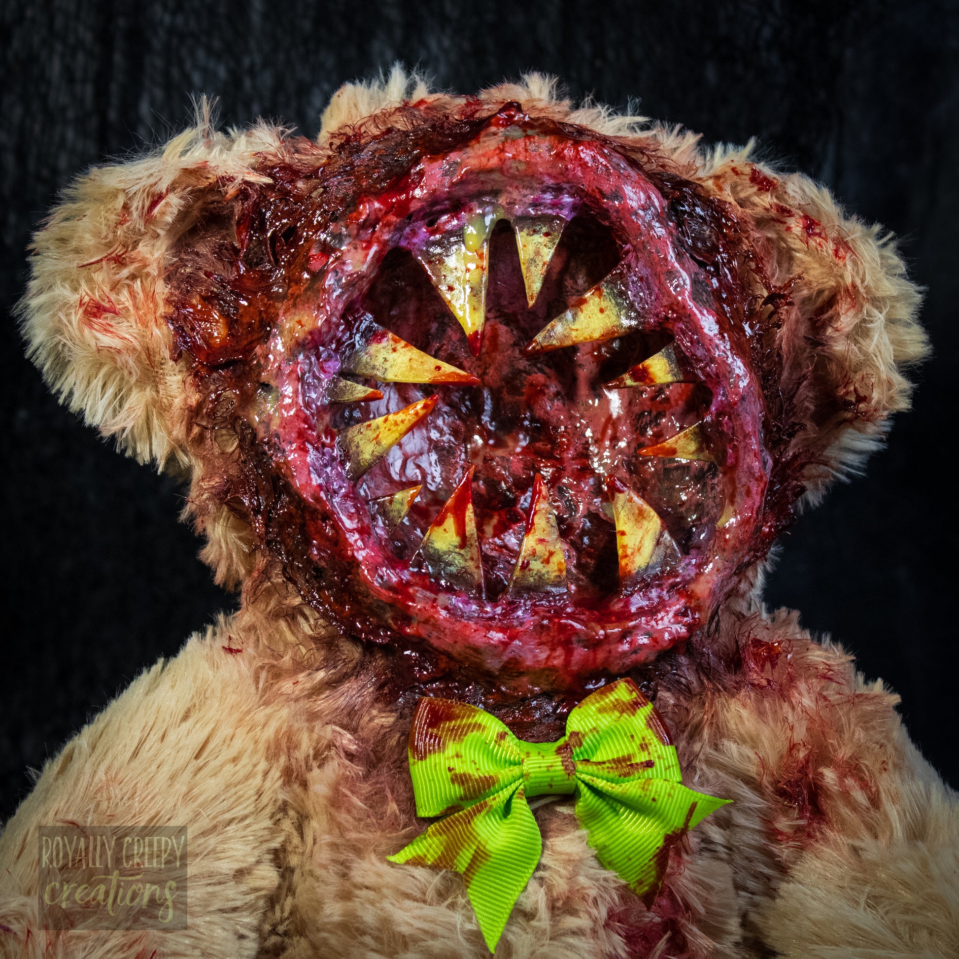 A close-up of a horror-themed plush teddy bear with a  big round grotesque mouth covering it's face. Big sharp yellow teeth and blood splatter detail. 