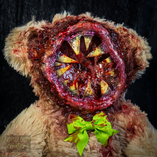A close-up of a horror-themed plush teddy bear with a  big round grotesque mouth covering it's face. Big sharp yellow teeth and blood splatter detail. 