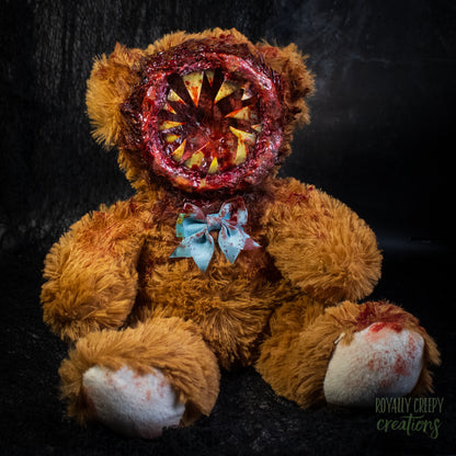 A horror-themed plush teddy bear with a round gory mouth with sharp yellow teeth covering the entire face. A blue bowtie at the neck, photographed against a dark textured background.