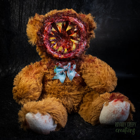 A horror-themed plush teddy bear with a round gory mouth with sharp yellow teeth covering the entire face. A blue bowtie at the neck, photographed against a dark textured background.