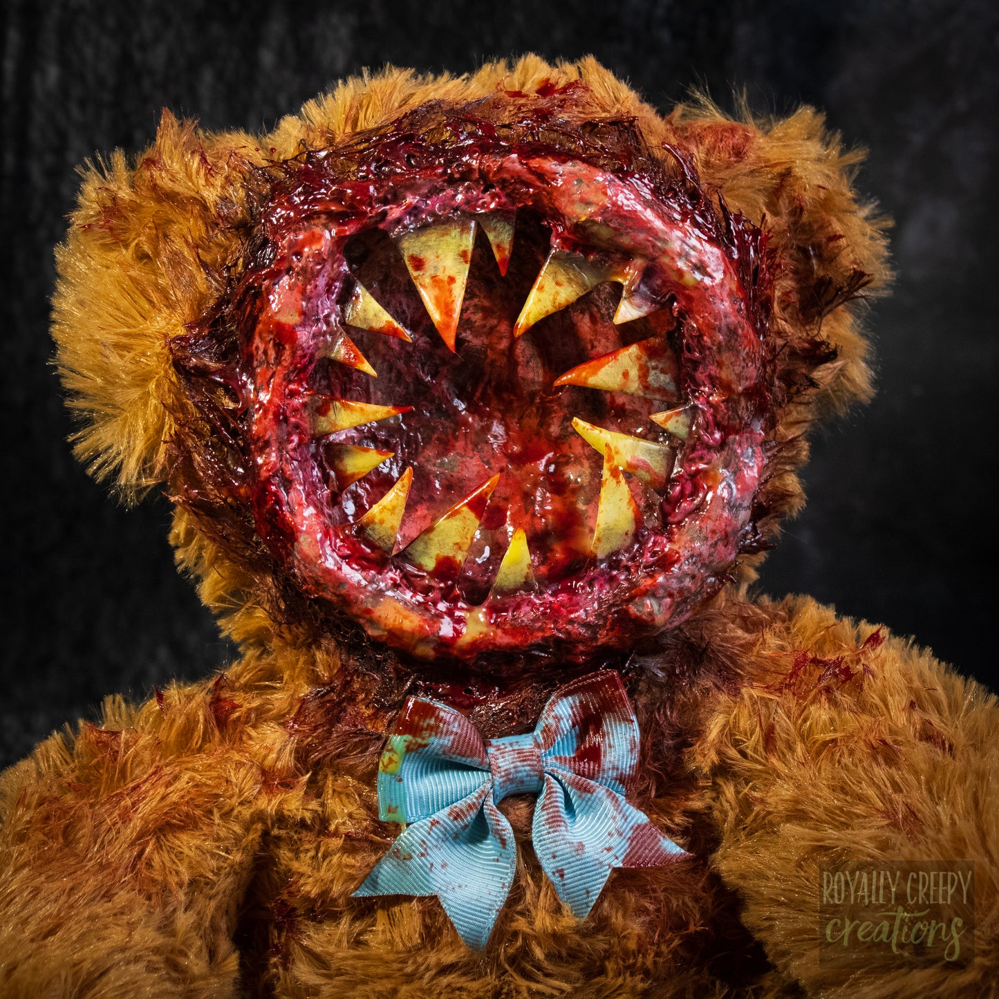 A close-up of a horror-themed plush teddy bear with a round gory mouth with sharp yellow teeth covering the entire face. A blue bowtie at the neck, photographed against a dark textured background.