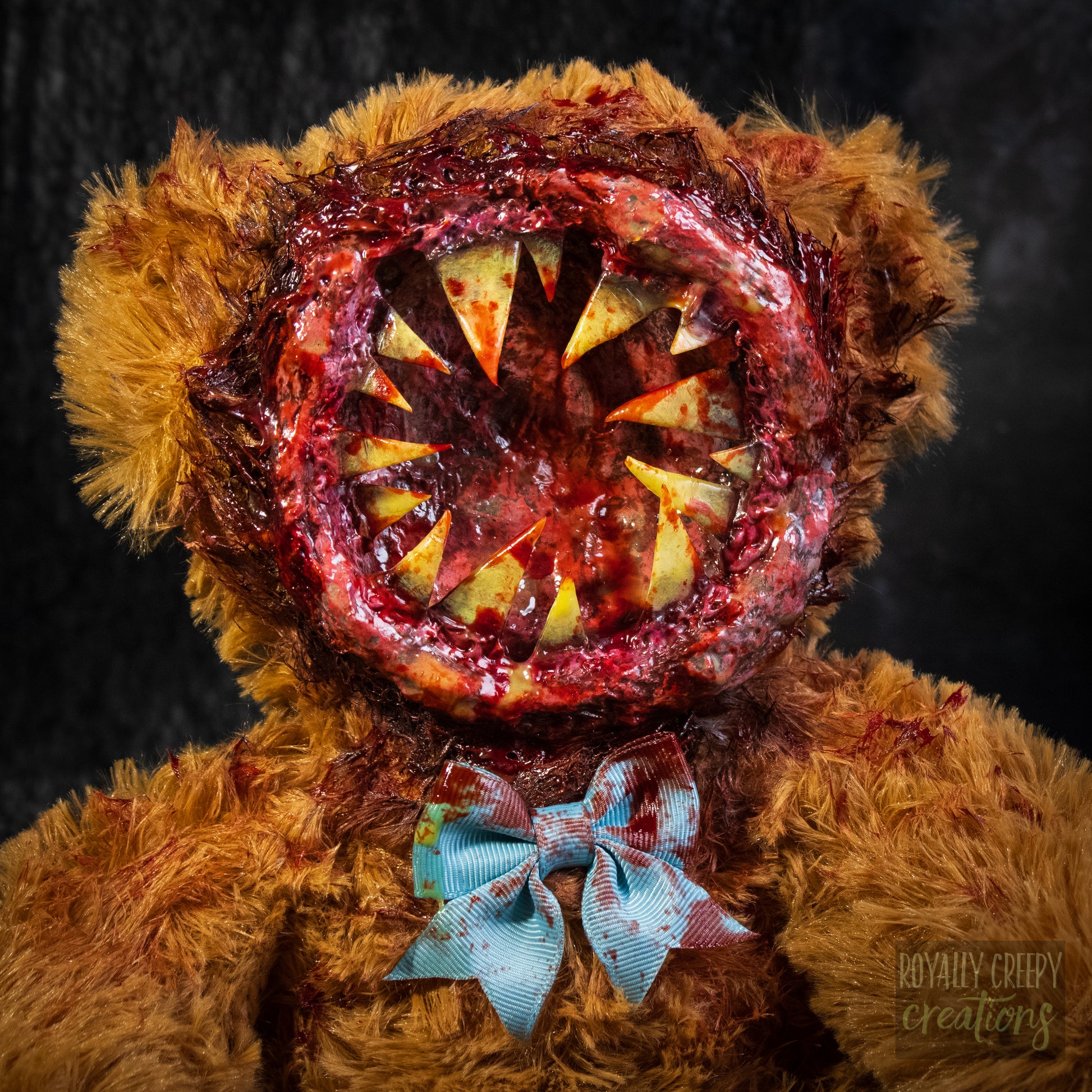 A close-up of a horror-themed plush teddy bear with a round gory mouth with sharp yellow teeth covering the entire face. A blue bowtie at the neck, photographed against a dark textured background.