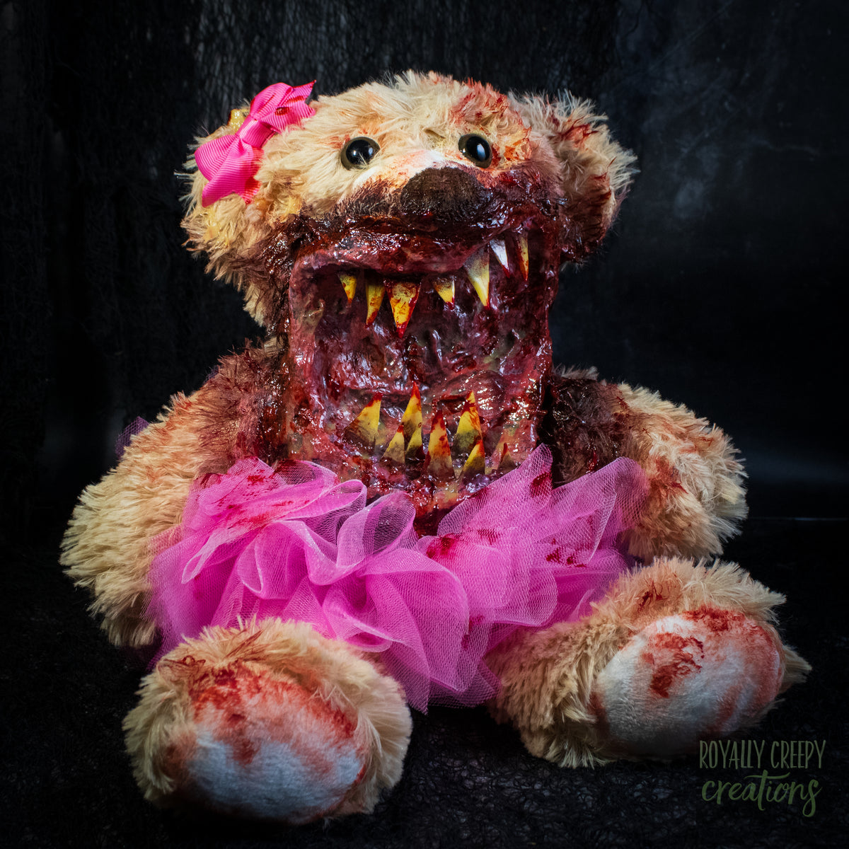A horror-themed plush teddy bear with a bloody, grotesque mouth showing sharp teeth, a bright pink tutu, and a pink bow on its head, set against a dark background.