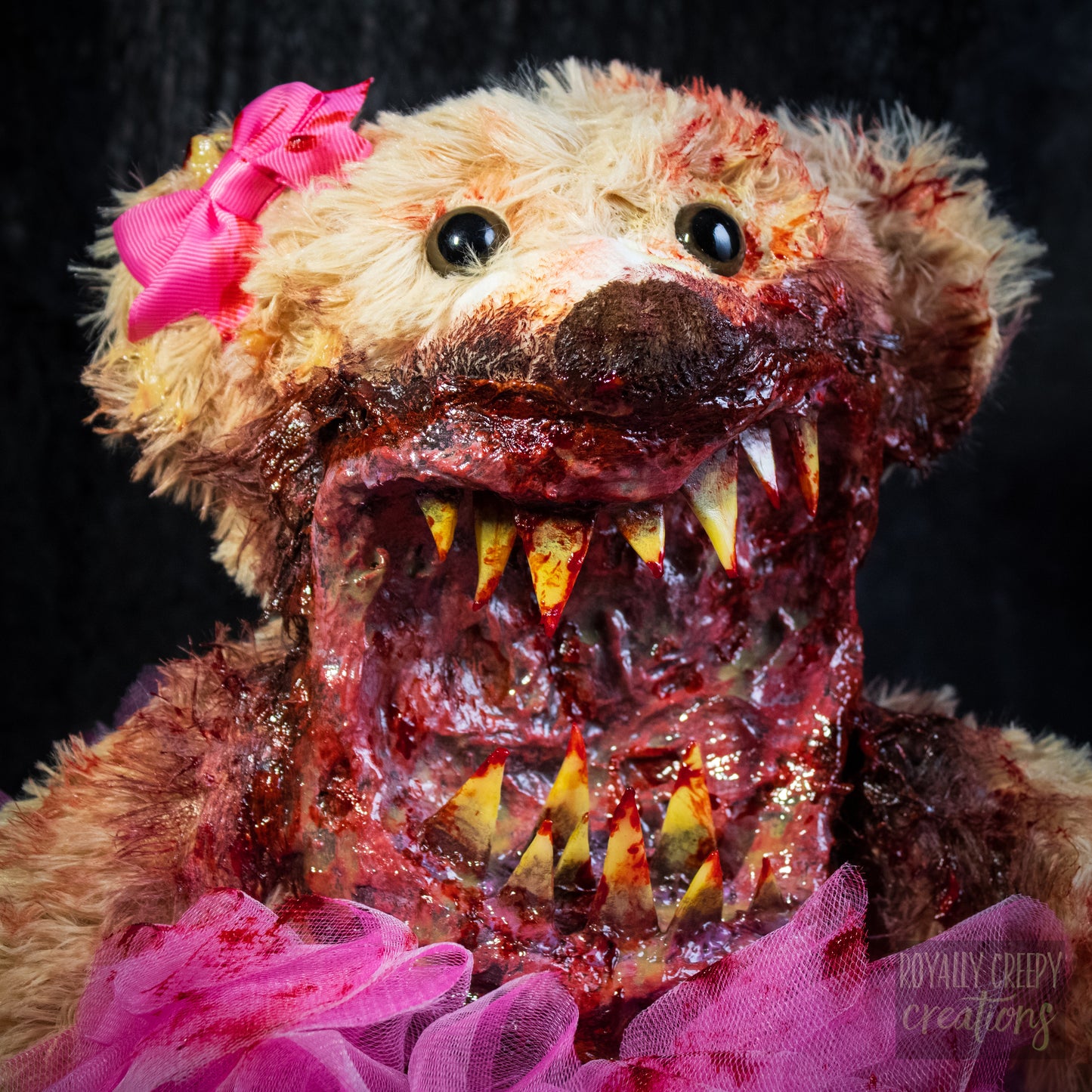 A close-up of a horror-themed plush teddy bear with a bloody, grotesque mouth showing sharp teeth, a bright pink tutu, and a pink bow on its head, set against a dark background.