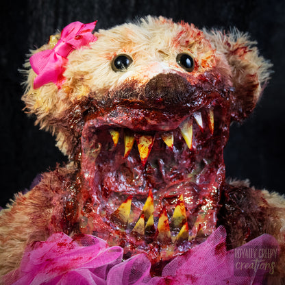 A close-up of a horror-themed plush teddy bear with a bloody, grotesque mouth showing sharp teeth, a bright pink tutu, and a pink bow on its head, set against a dark background.
