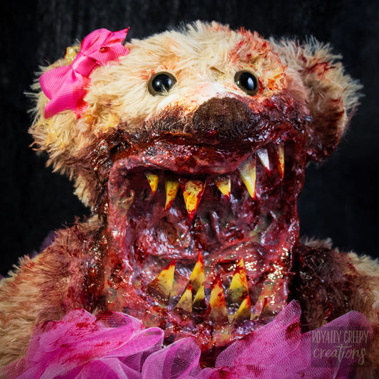 A close-up of a horror-themed plush teddy bear with a bloody, grotesque mouth showing sharp teeth, a bright pink tutu, and a pink bow on its head, set against a dark background.