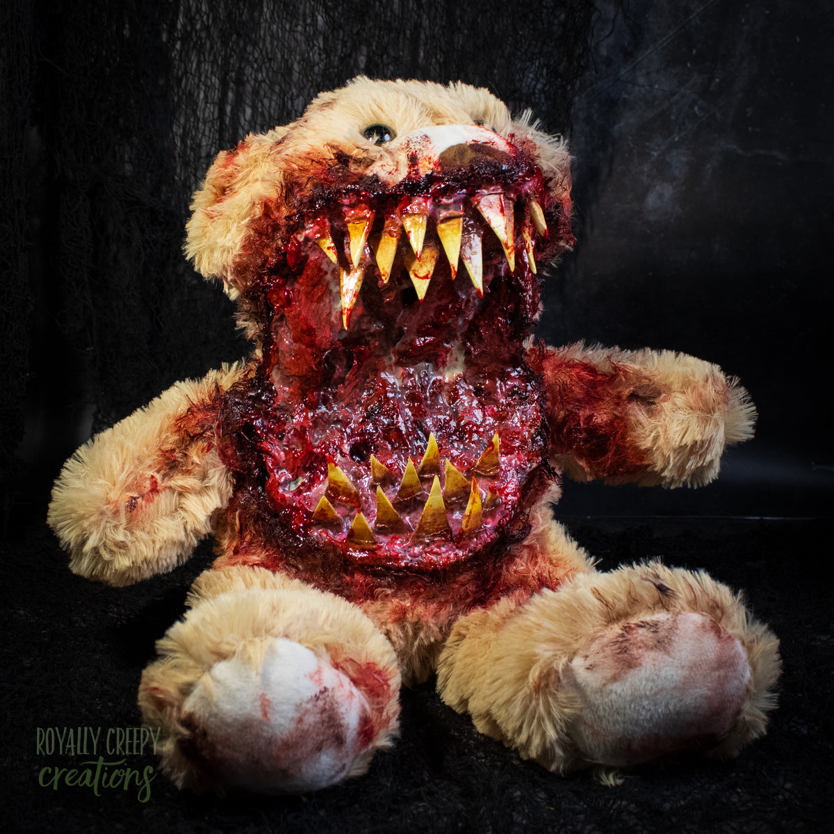 A horror-themed plush teddy bear with a grotesque, bloody monster mouth, sharp yellowed teeth, and exposed flesh-like detailing on the chest, set against a dark textured background.